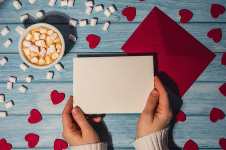 Female hands holding empty valentine card mock up red envelope with white cup of coffee and marshmallows on wooden blue background. Romantic Small hearts Valentine day. Blank paper card copy space for your text. Holiday morningの写真素材