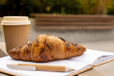 Fresh baked chocolate croissant on paper cup with lid for tea to go. Empty paper notebook. Freelancers place of work. Study and work. Remote business education. Making plans for next week year. Being mindful reducing stress and slowing-down. Take-out Take awayの写真素材