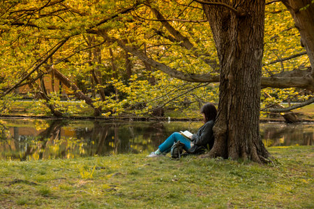 Attractive young woman reading book while sitting on grass in green public park. Springtime outdoors. Greenery unity with nature. Spend free time on open airの写真素材