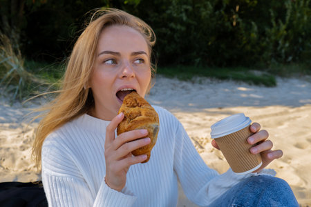 Happy young woman drinking coffee from paper cup takeaway food eating croissant on the beach sea...