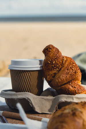 Tasty fresh croissants with coffee in paper cup on sandy beach sea coast background. Breakfast outdoors wellbeing concept. Take away food. Coffee alternativesの写真素材