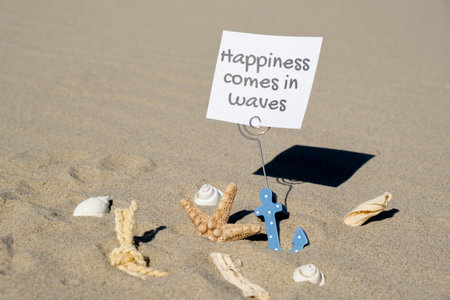 HAPPINESS COMES IN WAVES text on paper greeting card in anchor paper holder and starfish seashell summer vacation decor. Sandy beach sun coast. Holiday concept postcard. Getting away Travelの写真素材