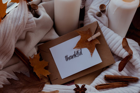 THANKFUL text greeting card concept Celebrating thanksgiving autumn holidays at cozy home on the windowsill Hygge aesthetic atmosphere Autumn leaves spices and candle on knitted white sweater in warm yellow lights. Still life.の写真素材