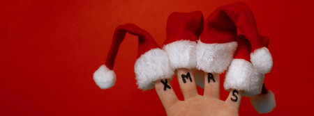 Fingers dressed in Santa-Claus red-white hats and word text XMAS on red background. Concept of...