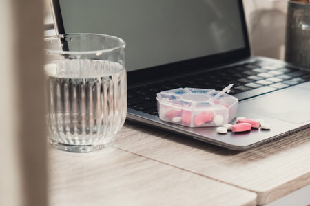 Organizer weekly shots with glass of water on workplace background and laptop Closeup of medical pill box with doses of tablets for daily take medicine with white pink drugs and capsules. Health care and diseases cureの写真素材