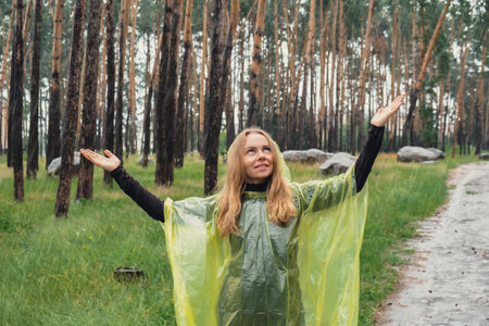 Happy blonde woman in yellow raincoat enjoying outdoors in forest. Rainy weather outside leisure activities in autumn time. Rainy seasonの写真素材