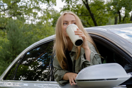 Blonde woman sticking head out of windshield car and drink coffee or tea from reusable mug. Young tourist explore local travel making candid real moments. Refuse reuse recycle zero waste concept. Responsible traveling reduce carbon footprintの写真素材
