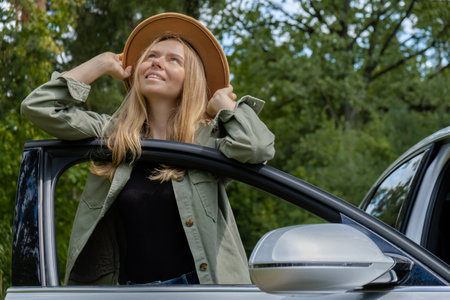 Blonde woman in hat staying next to car door. Young tourist explore local travel making candid real moments. True emotions expressions of getting away and refresh relaxの写真素材