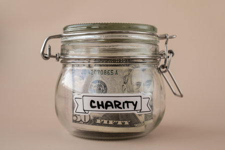 Saving Money In Glass Jar filled with Dollars banknotes. CHARITY transcription in front of jar. Managing personal finances extra income for future insecurity. Beige backgroundの写真素材
