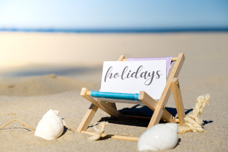 HOLIDAYS text on paper greeting card on background of beach chair lounge summer vacation decor. Sandy beach sun. Holiday concept postcard. Travelの写真素材