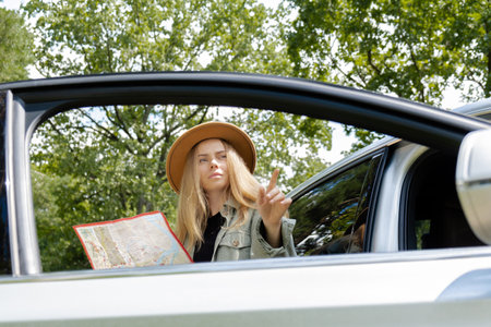 Blonde woman in hat staying next to car door checks the route on map to get to the destination....