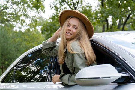 Smiling young woman in hat looking from car window. Local solo travel concept. Exited woman explore...