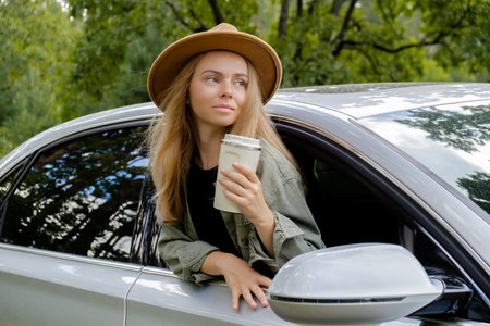 Smiling young woman looking from car window and drinking coffee or tea from reusable thermos cup....