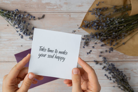 Female hands taking paper card note with text TAKE TIME TO MAKE YOUR SOUL HAPPY from violet envelope. Lavender flower. Top view, flat lay. Concept of mental spiritual health self care wellbeingの写真素材
