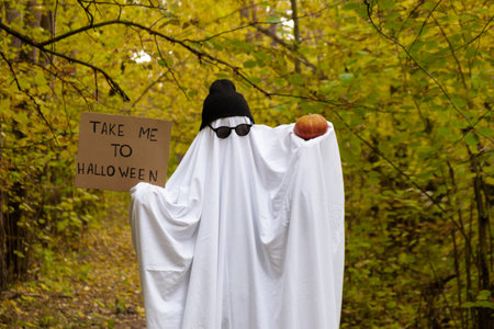 Man wearing ghost costume holding cardboard with text TAKE ME TO HALLOWEEN and pumpkin between orange yellow leaves in autumn forest for Halloween. Cool ghost creative holiday celebration fall decoration. Mysteriousの写真素材