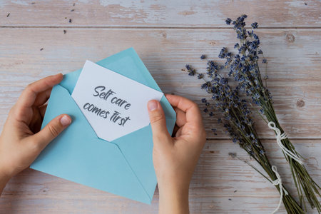 Female hands taking paper card note with text SELF CARE COMES FIRST from blue envelope. Lavender flower. Top view, flat lay. Concept of mental spiritual health self care wellbeingの写真素材