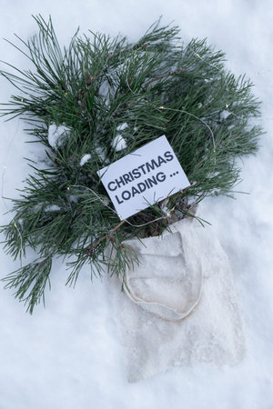 Paper note with text CHRISTMAS LOADING on Reusable shopping bag with fir spruce green branches in snowy outdoor winter forest background. Merry Christmas and new year concept. Natural bouquet greeting card copy spaceの写真素材