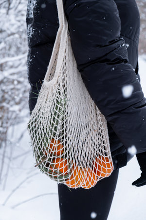 Unrecognizable woman holding Mesh shopping bag with tangerines in snowy winter forest outdoor. Go...