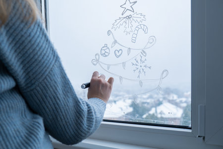 Creative enthusiastic unrecognizable woman in sweater paint window with white marker. Christmas tree decoration on big clear window. Snowy winter outside. Holiday preparationの写真素材