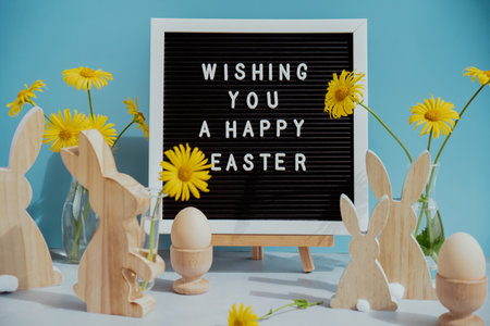 Phrase WISHING YOU A HAPPY EASTER traditional greeting to christian holiday. Festive mood composition with bunnies and yellow flowers on pastel blue background. Sustainable natural materialsの写真素材