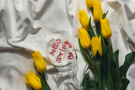 Hello SPRING message on wine glass with yellow fresh tulips on white bed with trendy deep shadows....