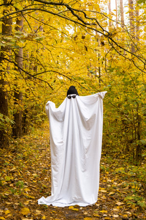 Happy Halloween holiday. Cute unrecognizable person dancing in ghost costume on fall forest background. Creative costume with white and black cap hat and glassesの写真素材