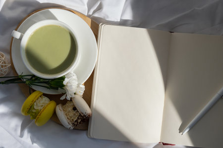 White pages of notebook copy space Sweet yellow macaron dessert Cup of green plant based matcha tea latte in cozy morning bed atmosphere. Creative holiday romantic idea. Herbal prebiotic fiber superfood beverage antioxidant-rich immunity-boostingの写真素材