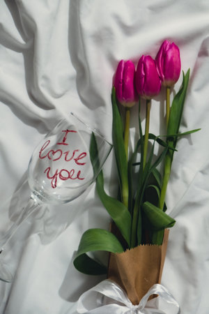 Bouquet of pink tulips on bed. I love you written text on wine glass. Romantic morning scene. Greeting card Holiday flowers springtimeの写真素材