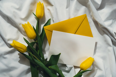 Mock up of white empty envelope greeting card with yellow fresh tulips on white bed with trendy deep shadows. Spring flowers in yellow colors. Romantic postcard wallpaper designの写真素材