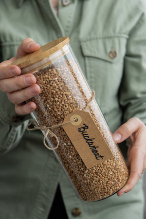 Unrecognizable woman holding refillable glass jar with raw buckwheat porridge. Refill product format eco-friendly living. Filling reusable container Whole spelt grain species. High protein fiber-rich vegan plant based food ingredient. Gluten free superfood healthy nutritional dietaryの写真素材