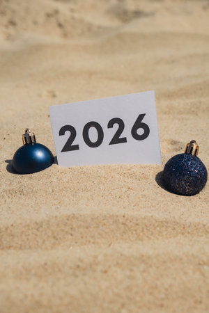 2026 text on paper greeting card on background sandy beach sun coast. Christmas balls New Year New...