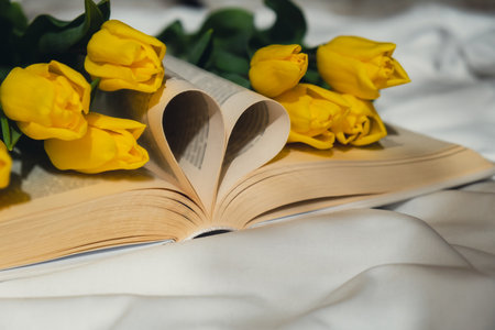 Book pages in shape of heart symbol love with bouquet of pink tulips on messy bed. Romantic morning...