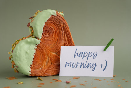 HAPPY MORNING caption on paper note love symbol Heart shaped pistachio croissant on green background with copy space. Puff pastry trendy sweet dessert bakery. Creamy dough experimentの写真素材