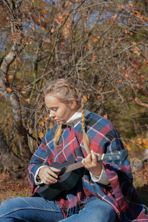 Portrait of smiling caucasian girl plays ukulele in autumnal park. Young woman plays guitar musical instrument outside in nature fall time. Audio music healingの写真素材