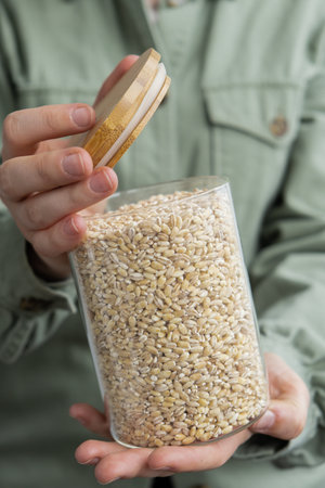 Unrecognizable woman holding refillable glass jar with raw pearl barley porridge. Refill product format eco-friendly living. Filling reusable container Whole spelt grain species. High protein fiber-rich vegan plant based food ingredient. Gluten free superfood healthy nutritional dietaryの写真素材