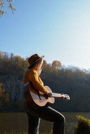 Blonde woman playing string guitar outdoors in autumn forest. Concept of sound therapy, mental health and wellness rituals. Calmness tranquility audio-sensory practices. Aura farming energyの写真素材