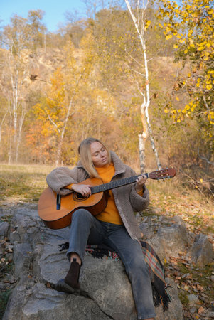 Blonde woman playing string guitar outdoors in autumn forest. Concept of sound therapy, mental health and wellness rituals. Calmness tranquility audio-sensory practices. Aura farming energyの写真素材