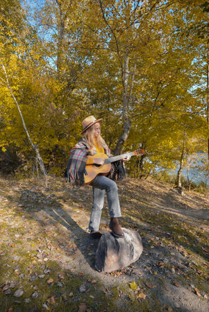 Beautiful blonde girl playing acoustic guitar and singing relaxing enjoying outdoors on sunny autumn...