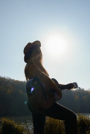 Beautiful blonde girl playing acoustic guitar and singing relaxing enjoying outdoors on sunny autumn day. Hippie woman in boho style learning and practicing playing musical instrument. Fall leavesの写真素材