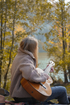 Blonde woman playing string guitar outdoors in autumn forest. Concept of sound therapy, mental...