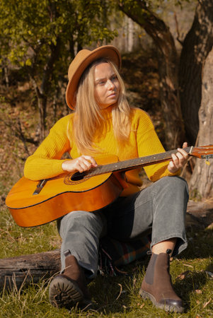 Blonde woman playing guitar outdoors in autumn forest. Concept of sound therapy, mental health and...
