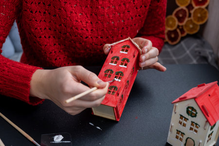 Sustainableminded Christmas cozy atmospheric festive decor in modern scandinavian home handmade decoration. Unrecognizable woman in red sweater painting craft wooden houses. Seasonal activity for kidsの写真素材