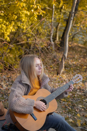 Blonde woman playing string guitar outdoors in autumn forest. Concept of sound therapy, mental health and wellness rituals. Calmness tranquility audio-sensory practices. Aura farming energyの写真素材