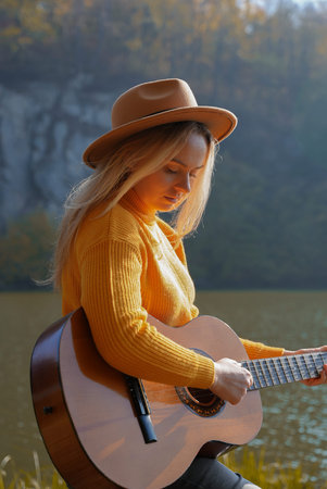 Blonde woman playing string guitar outdoors in autumn forest. Concept of sound therapy, mental...