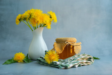 Golden dandelion jelly presented in glass jar. Fresh yellow dandelion flowers surround the jar, emphasizing natural ingredients and traditional homemade preparation. Concept of homemade food