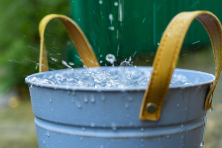 Rainwater Harvesting System Collecting Roof Runoff into Bucket for Sustainable Water Conservation. Rainwater flows from a roof gutter and drainage system into a metal bucket for collection and future use