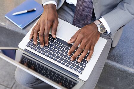 Close-up of African businessman sitting with laptop computer on his knees and typing on it outdoorsの写真素材