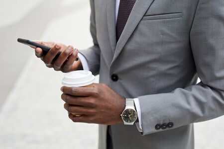 Close-up of African businessman in suit holding cup of coffee and using mobile phone for his online work outdoorsの写真素材