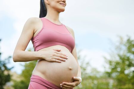 Young pregnant woman in pink clothing standing and touching her belly outdoors she expecting her...