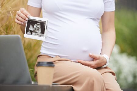 Close-up of pregnant woman holding ultrasound of her baby while sitting on the bench with laptop and...
